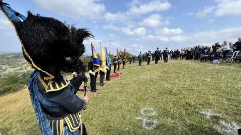 A group of people watching a military service on on Cleeve Hill, including several people in military uniforms and a bagpiper, to remember seven men who died 