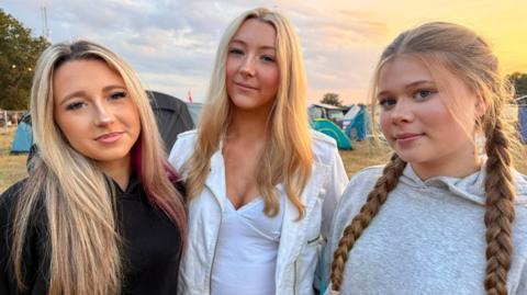 All the girls have long blonde hair. Caitlin wears a black jumper, Abi in the middle wears a white jacket and Libby has hair in plaits and is wearing a grey hoodie.