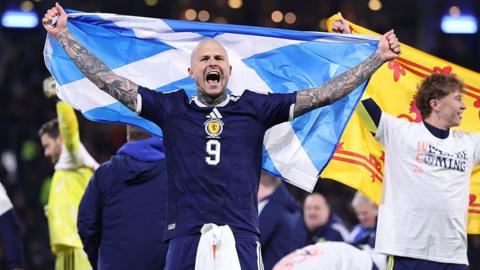 Scotland striker Lyndon Dykes celebrates getting to the World Cup. He is in a Scotland number nine shirt and has a Scotland flag held up behind him.