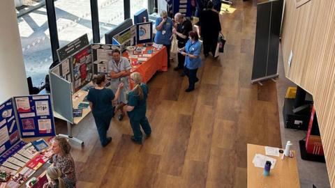 An aerial view of a previous event where men and women dressed in medical outfits are standing chatting next to tables filled with leaflets and information for the public. Other people are seen looking at some of the leaflets and walking around what looks likes a leisure/health centre with laminate flooring and wood panelling.