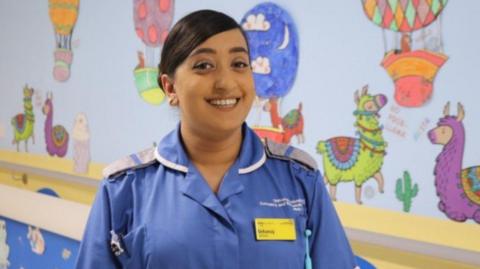 A woman wearing a nurses uniform stands in front of a colourful wall.