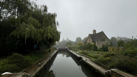 A canal under grey clouds in Iffley with a tree's branches handing over the water and stone building in the background. 