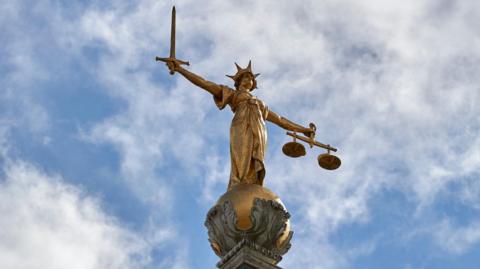 Lady Justice, a 12ft high, gold leaf statue pictured on top of the dome of the Central Criminal Court, The Old Bailey, in central London. The sky is blue with some fluffy white clouds in the background.