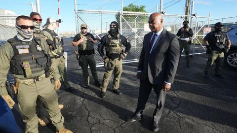 Mayor Baraka, seen visiting the same facility on Wednesday. He is a black man, and wears a dark suit. Armed officers wearing masks gather around him