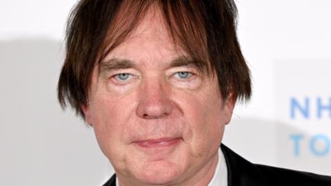 A man with dark hair that has been brushed to either side of his face is pictured looking expressionless in front of a white background. He is clean shaven and has greyish blue eyes.