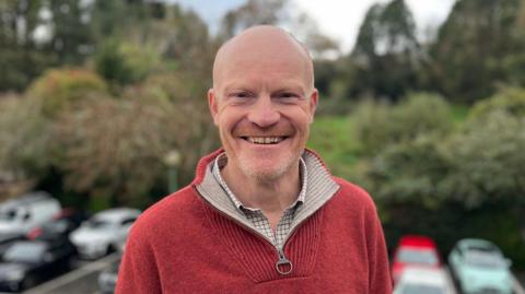 Deputy Gavin St Pier - A man with a bald head and a ginger and grey beard smiling at the camera in a car park. He is wearing a red jumper. 