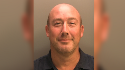 A mug shot of a man looking into the camera. He is bald with light stubble. He is wearing a dark blue collared shirt. 