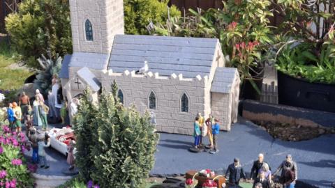 A model of a white church with a grey slate roof. There are tiny figurines around the church.