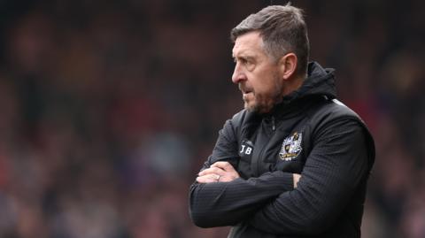 Port Vale boss Jon Brady crosses his arms and looks on from the touchline 