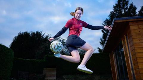 Isabel Wilkins wearing white trainers, black shorts and a red football jersey over a black long-sleeved top. She has her light brown hair tied in a ponytail and is jumping high in the air doing a trick with the football. She appears to be in a back garden at dusk, next to a wooden shed and some trees. The photographer has used a bright flash and the background is dark.