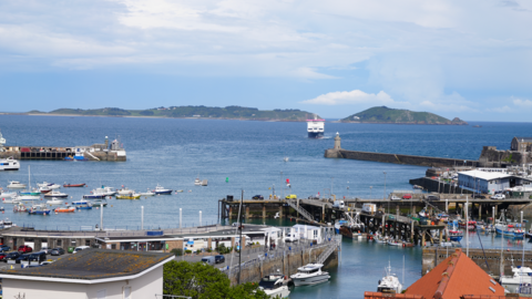 A coastal harbor scene featuring numerous boats and yachts docked along piers. A large ferry or ship is visible in the distance, approaching the harbor. Surrounding the harbor are buildings and docks, with hills or small islands in the background beneath a partly cloudy sky.

