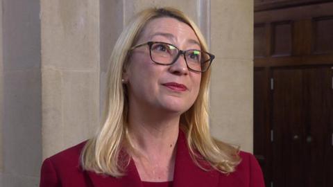 Jess Asato during an interview. She looks to the right of the camera. She has shoulder length blonde hair, glasses and is wearing a red blazer.