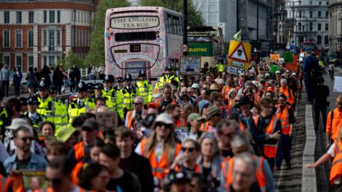 A row of several dozen protesters all wearing orange high visibility vests on a London street with London busses and police officers in the background