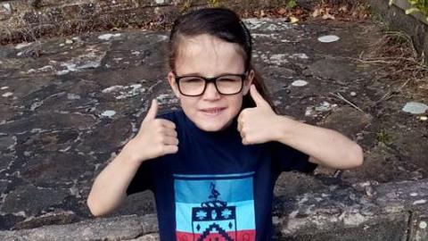 A smiling Millie Gribble in front of a stone wall holding her two thumbs up. She has long dark hair tied back in two bunches with a hair clip at the side. She is wearing a navy football t-shirt.