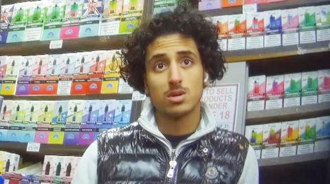 A young man called Akwa, captured by secret filming, stands inside a shop, in front of shelves with rows of colourful packaging for vapes. He wears a shiny black bodywarmer with a grey top underneath, he has curly short dark hair and he looks to be in conversation.