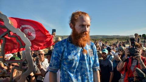 Russ Cook wearing a blue England shirt, with a long ginger beard stood on a platform. There is a crown around him, and a red Tunisia flag in the background. 