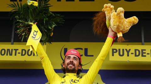 Ben Healy celebrates on the podium wearing the yellow jersey after stage six of the Tour De France. He is holding a stuffed animal in his left hand and flowers in his right. 