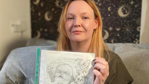 Kelly has brown hair and is holding a sketch of her son Christopher. There is artistic wallpaper behind her.