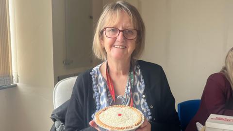 Pat Limb - who has short blonde hair - is holding a Manchester Tart. 