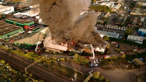 An aerial drone photograph above an industrial fire in Witham. Black smoke is billowing into the air and orange flames are coming off the roof. There are numerous fire engines at the scene with aerial platforms.