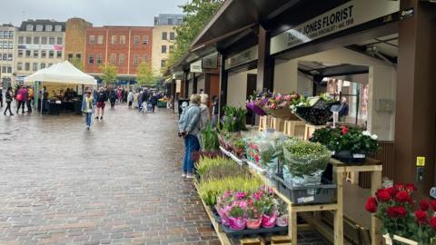 Brown and cream market stall with buckets of flowers including red roses on the right.  Other stalls are visible in the distance.  Shoppers are looking at the produce. The paving shows signs of rain.