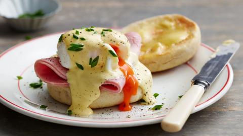 A plate with a single serving of eggs Benedict: a toasted English muffin, one half buttered and one half topped with slices of ham, a poached egg, and hollandaise sauce.