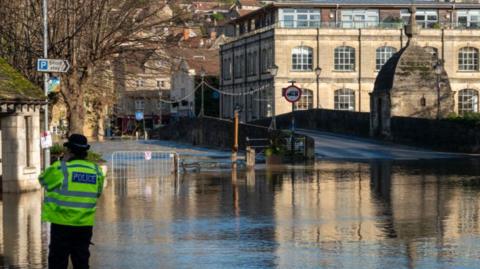 Flood water surrounds the town bridge which spans the River Avon which has burst its banks and flooded nearby riverside properties on November 25, 2024 in Bradford on Avon, England.  There's a police officer wearing a florescent jacket standing by the edge of a flooded road. It's sunny.