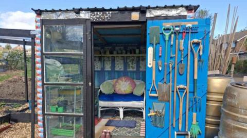 A shed with glass doors, a small sofa chair and tools on the wall. It is in the middle of an allotment.