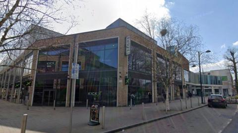 A large building with glass windows stretching the length of the facade. A street with bollards and a car parked on it stretches in front of it.