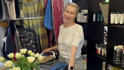 A woman stands smiling behind an ironing board. There are racks of clothes behind her and a vase of white roses to one side. She is holding an iron in one hand and a pair of jeans with the other. 