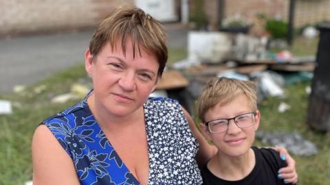 Jennifer with her arm around her son 12-year-old Nicholas outside their home, surrounded by debris and rubbish. 