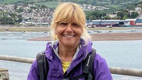 A woman with blonde hair standing by a railing with an estuary behind her. She is smiling at the camera and wearing a purple waterproof jacket and black rucksack. There are boats on the water behind her, and houses rising up a hillside.
