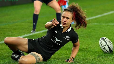 Ruby Tui, in all black New Zealand kit, points towards the camera as she slides along the ground, with ball on ground to right of picture