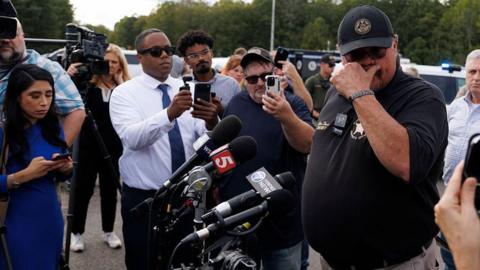 Sheriff in black polo and black cap holds hand up to face and looks away, amid microphones and reporters holding cameras and phones