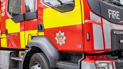 The side of a red fire engine, showing the door, wheel and the red and yellow markings.