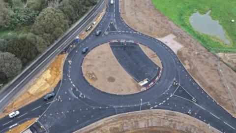 An aerial image of a roundabout under construction. It is surrounded by trees, greenery, and mud.