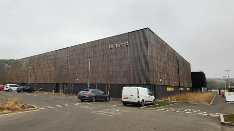 The Graham Sports Centre at Maiden Castle, Durham University Sports Complex. It is a large industrial unit that has brown panelled cladding. there are several cars and vans parked outside. It is a grey cloudy day.