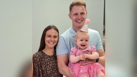 Rebecca and Harry Foster with their baby daughter, Olivia. Rebecca has long dark hair and is looking directly at the camera and smiling. She is standing next to Harry. He has blonde short hair and is wearing a light blue polo shirt. He is holding Olivia. She has blonde short hair and is wearing a pink and white stripe top with a pointed party hat.