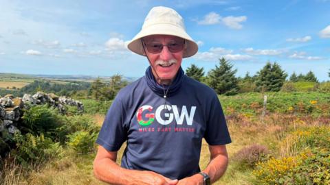 John Parry stands smiling at the camera, with a view of north Pembrokeshire and the Pembrokeshire coast behind him. John is wearing a 'grandad's great walk' fundraiser logo t-shirt, with sunglasses and a sun hat. He wears sunglasses and a white moustache. 