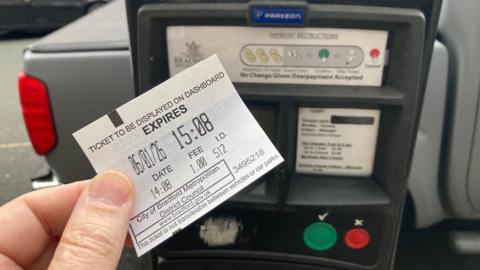 A man's hand holding a white parking ticket with black type in front of ticket machine