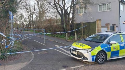 A police car partially blocks the entrance to a cycle path, which has also been cordoned off with blue and white tape. Trees line both sides of the path.