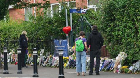 A few people look down at floral tributes placed outside Minehead Middle School. There are lots of bunches of flowers places along the pavement, next to the fence. 