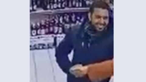 A CCTV image of a man in a dark jacket smiling as he shakes another man's hand inside a business at Cotham in Bristol