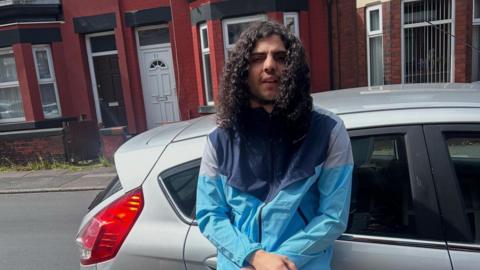 Kade Hassan-Hughes is pictured standing next to a silver-coloured car which is parked in front of a row of red brick terraced houses. He has shoulder-length, curly, dark hair as well as facial hair. He is wearing a light blue jacket.