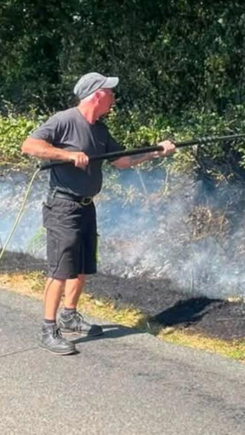 A window cleaner wearing a black T-shirt, shorts and boots uses a window brush to put out a fire on a grass verge at the side of a road