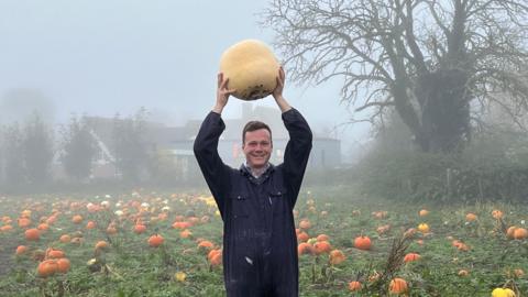 A man standing in a field full of pumpkins raises a large pumpkin above his head. He is wearing blue overalls.