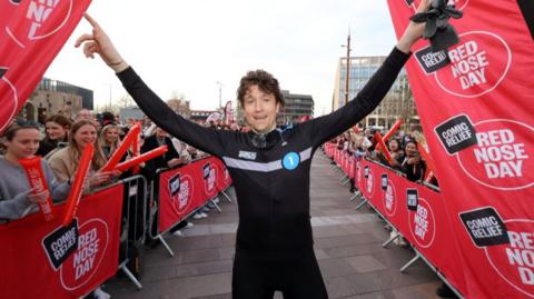 Greg James with his arms out stretched next to Red Nose Day flags and crowds of spectators.
