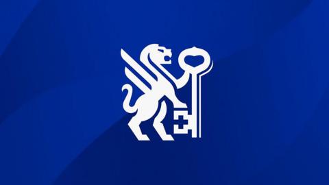 A crest consisting of a white winged lion standing up and resting its front leg on a key. It is on a dark blue background