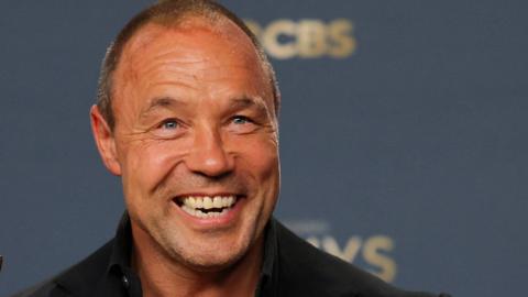 Stephen Graham, with a shaved head, grins at the camera in front of a blue wall
