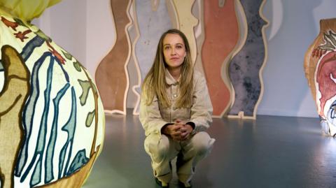 Bethany Williams wears a beige jacket and trousers, she has brown/ blonde hair and crouches in an exhibition which has colourful lights and arching coloured panels in the background.